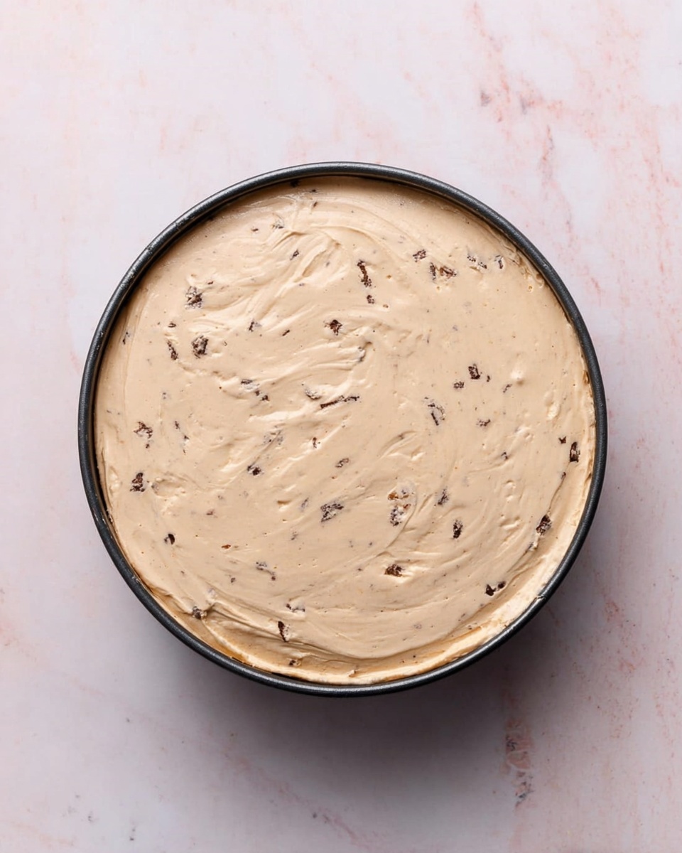 The image shows a round dessert topped with many pieces of honeycomb candy covered in chocolate. The candy pieces vary in size, some are whole chunks with a yellow honeycomb center surrounded by a chocolate coating, and others are crushed into small broken bits scattered across the surface. These candy pieces cover the entire top layer of the dessert on a smooth light brown base. The dessert sits on a white plate, placed on a white marbled surface. Photo taken with an iphone --ar 4:5 --v 7