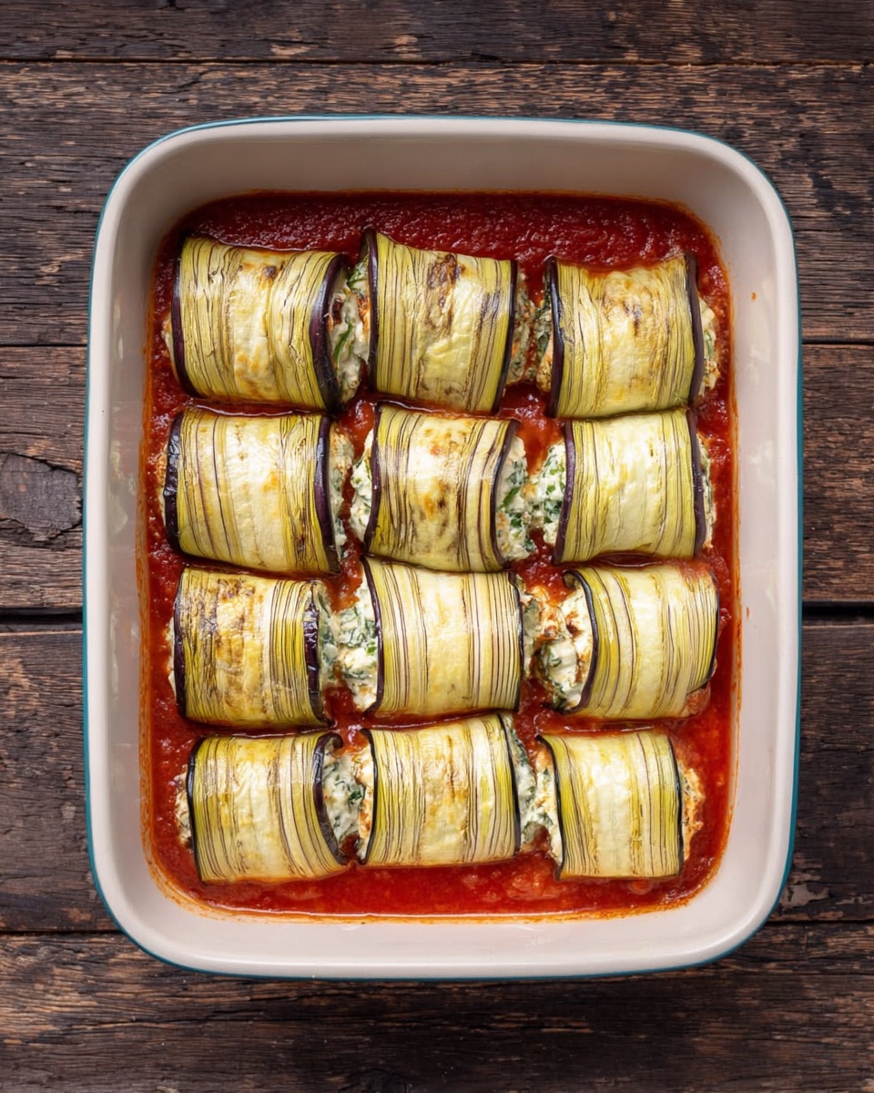 The image shows a white rectangular baking dish filled with eight rolled eggplant slices. Each roll is stuffed and topped with melted cheese that is golden brown in spots, sprinkled with finely chopped green herbs. The eggplant skin is dark purple, peeking out from under the cheese layer. Beneath the rolls, there is a rich red tomato sauce that has bubbled up around the edges, with some baked sauce bits visible. The dish is placed on a white marbled surface, and the background shows part of a stovetop blurred out. Photo taken with an iphone --ar 4:5 --v 7