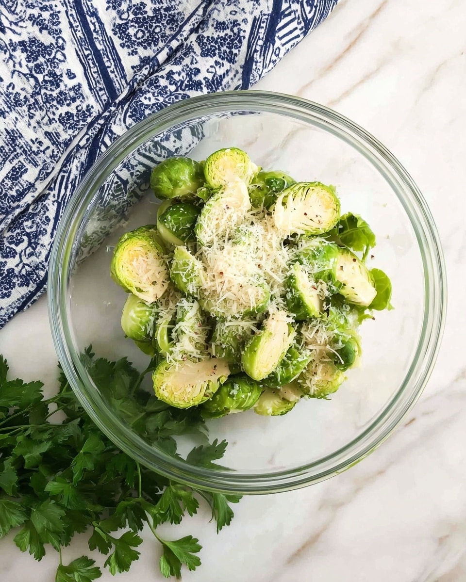 A clear glass bowl holds halved bright green Brussels sprouts arranged in a loose layer, topped with a sprinkle of fine white grated cheese and small black pepper specks. The bowl sits on a white marbled surface with a folded blue and white patterned cloth and fresh green parsley next to it. Photo taken with an iphone --ar 4:5 --v 7
