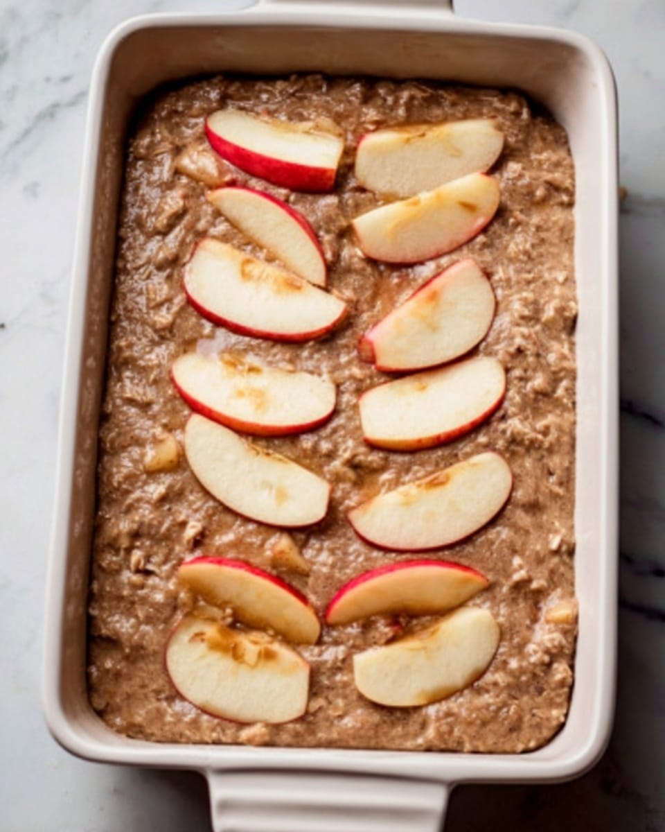 The image shows a white rectangular baking dish filled with a thick brown batter that contains small pieces of chopped apples mixed throughout. On top, there are seven sets of thin apple slices arranged in small, neat half circle groups, spaced evenly across the surface. The layers include the light brown batter with rough texture and visible apple chunks beneath the smooth, shiny apple slice toppings that create a gentle pop of red and cream colors. The dish is placed on a white marbled surface, giving a clean and bright look. photo taken with an iphone --ar 4:5 --v 7