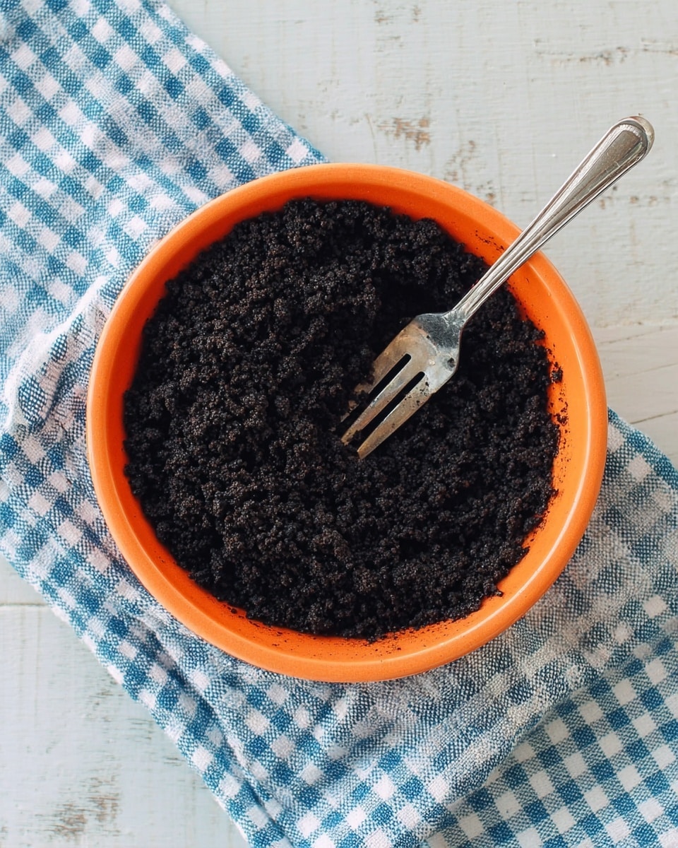 An orange round bowl filled with a single layer of dark black crumbly mixture that has a rough texture. A small silver fork is resting on the right side of the bowl, partly buried in the crumbs. The bowl is placed on a white and blue checkered cloth, which sits on a white marbled surface. Photo taken with an iphone --ar 4:5 --v 7