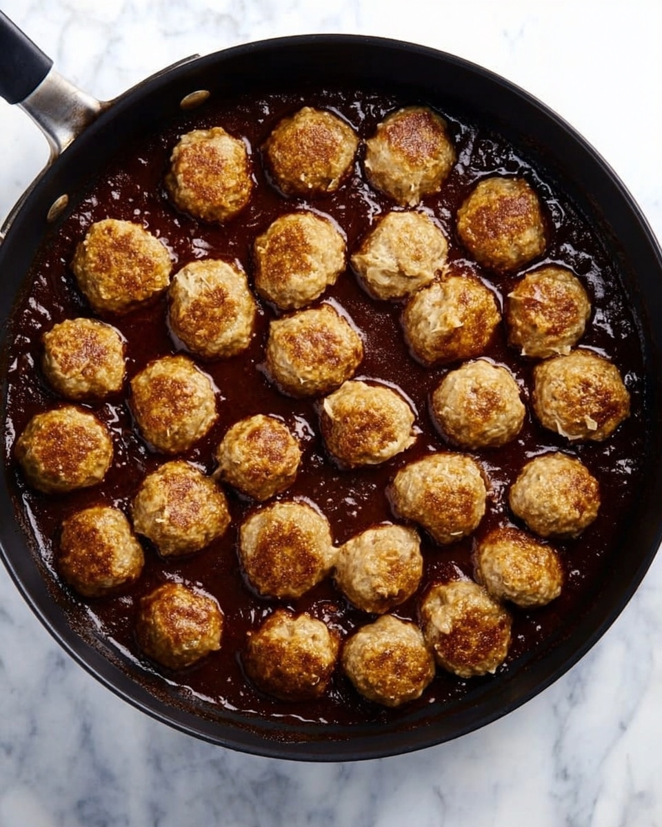 A black pan filled with many round meatballs all covered in a shiny dark brown sauce. The meatballs are evenly spaced and sit in a thick layer of sauce that has a glossy texture, making them look coated and juicy. The pan itself is on a white marbled surface, and no other items are visible around it. The lighting is natural and shows the sauce reflecting light, emphasizing its rich color and texture. photo taken with an iphone --ar 4:5 --v 7