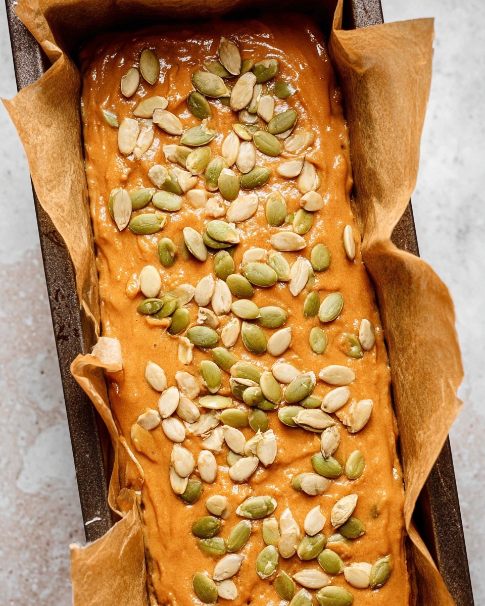 The image shows a close-up of a loaf pan lined with parchment paper filled with an orange-brown batter, smooth with some slight ridges. The top of the batter is scattered with many light green and pale pumpkin seeds evenly spread across the surface. The edges of the pan reveal the parchment paper folding upwards. The whole scene is set on a white marbled texture. photo taken with an iphone --ar 4:5 --v 7