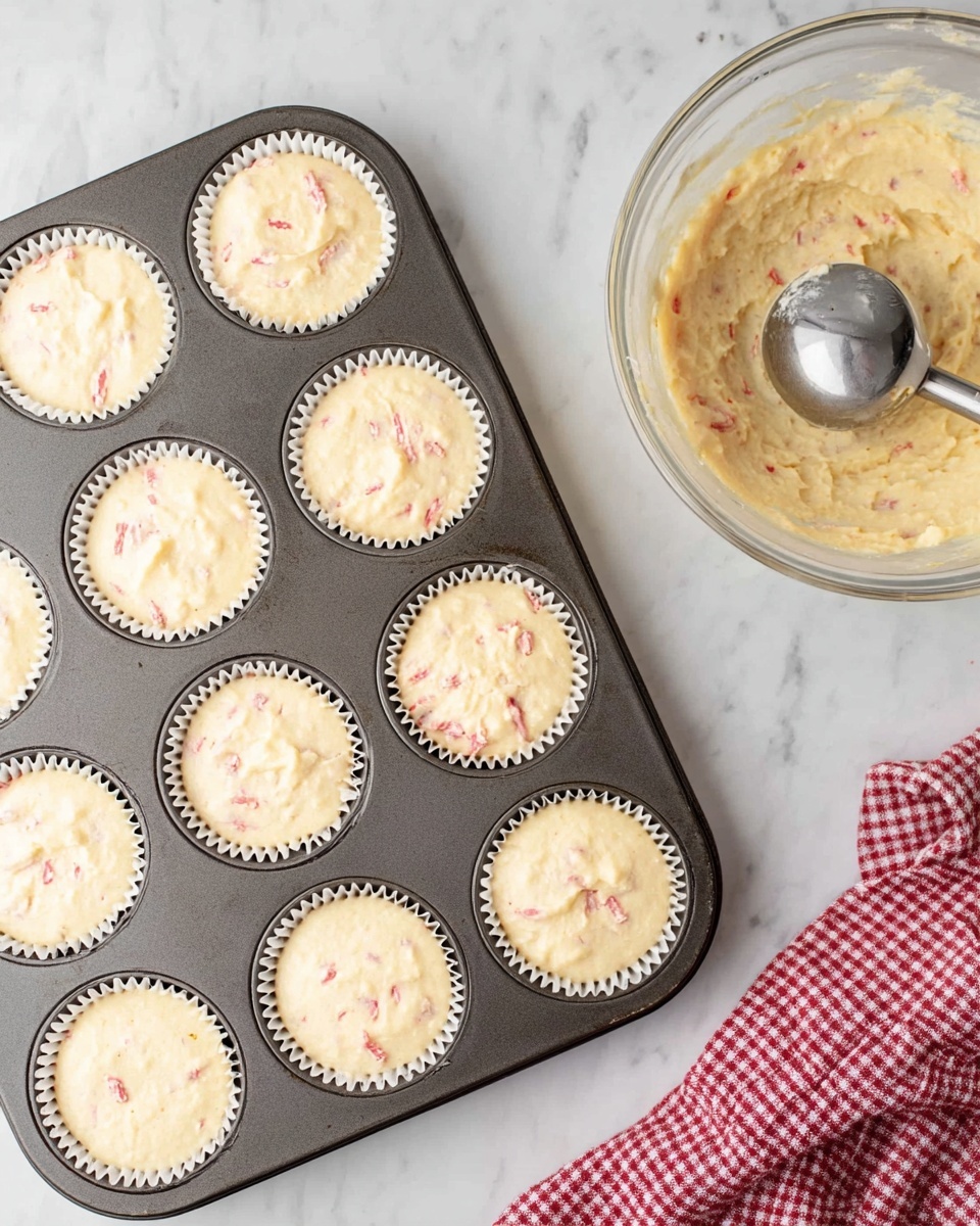 The image shows four small cheesecakes on a white marbled surface. Each cheesecake has three layers: a thick brown crumbly base, a creamy white middle layer with red bits inside, and a tall swirl of white whipped cream on top. The whipped cream is sprinkled with crushed red candy pieces. Around the cheesecakes, there are whole and crushed red and white peppermint candies, with a small bunch of green mint leaves to the side. The photo is taken with an iphone --ar 4:5 --v 7