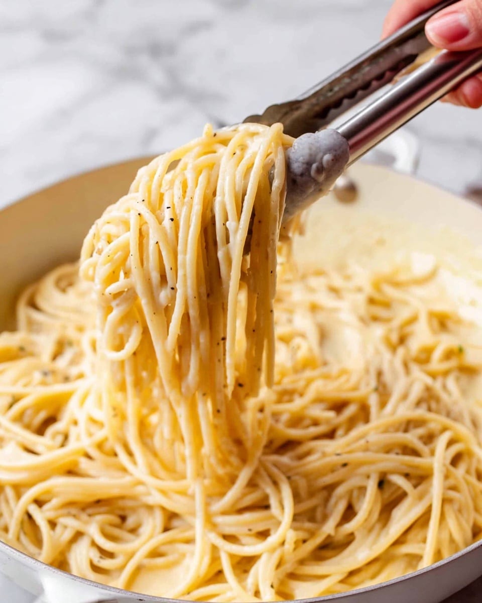 The image shows a close-up of cooked spaghetti being lifted by silver tongs over a white pan filled with more spaghetti. The spaghetti strands are pale yellow and smooth, coated lightly with a glossy, creamy sauce with small specks of seasoning. A woman's hand is holding the tongs from the bottom left of the photo. The background is a white marbled surface. photo taken with an iphone --ar 4:5 --v 7