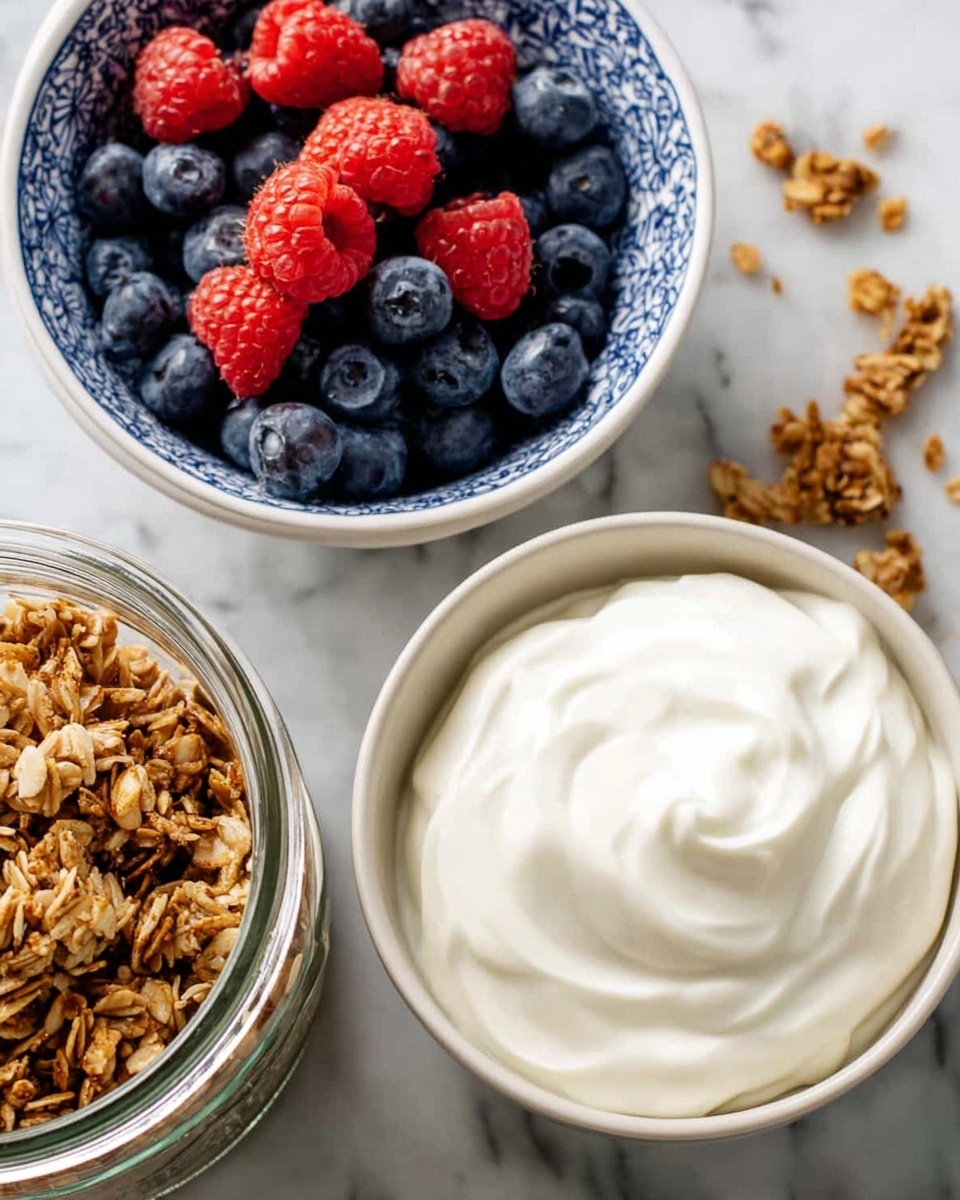 A white ceramic bowl filled with smooth, creamy yogurt sits slightly to the right in the image, showing soft swirls on top. To the left of the yogurt, there is a white bowl with blue patterns filled with fresh blueberries and bright red raspberries, showing a mix of round and bumpy textures. Below these bowls, a clear glass jar holds a golden brown granola mix with oats and a crunchy look. These items are placed on a white marbled surface with a few stray granola pieces scattered around. Photo taken with an iphone --ar 4:5 --v 7