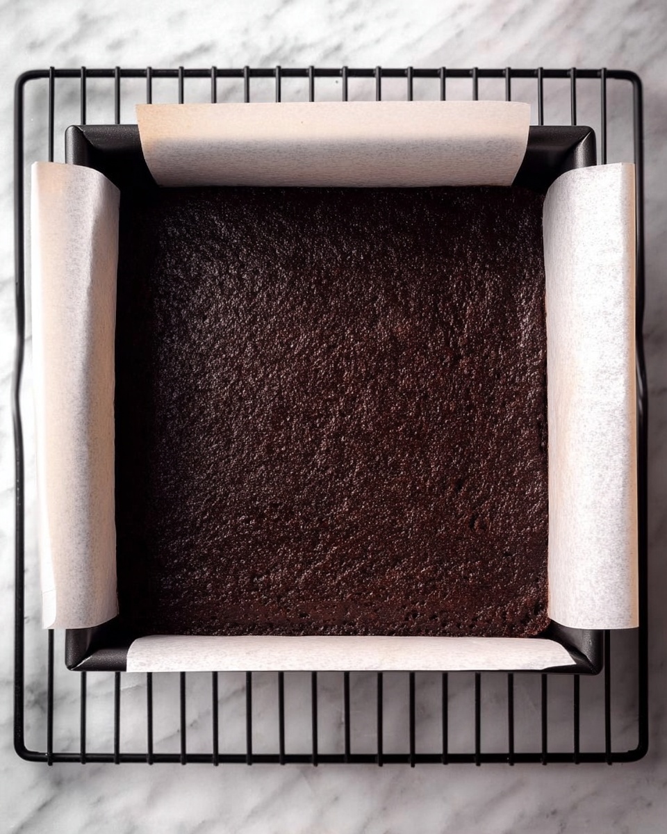 The image shows a square baking pan lined with white parchment paper that extends beyond the edges of the pan. Inside the pan is a dark brown baked cake layer with a slightly rough and uneven texture on the surface. The pan rests on a black cooling rack placed on a white marbled surface. photo taken with an iphone --ar 4:5 --v 7
