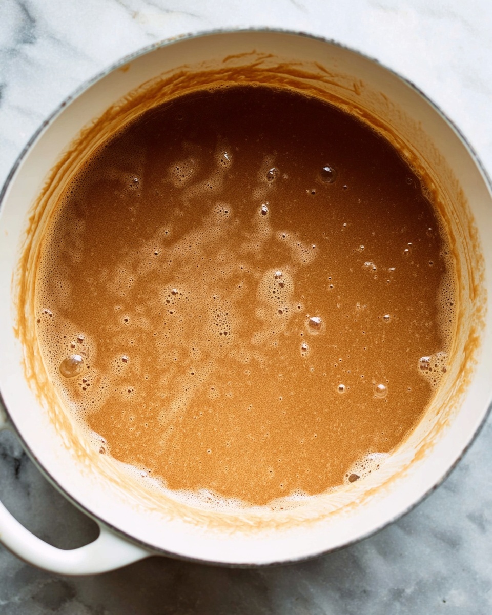 A white pot filled with a thick, light brown liquid that looks creamy and smooth, with bubbles and small lumps on the surface. The liquid nearly reaches the top edge of the pot, and there are some streaks of the same light brown color on the inner sides of the pot. The pot sits on a white marbled surface. photo taken with an iphone --ar 4:5 --v 7