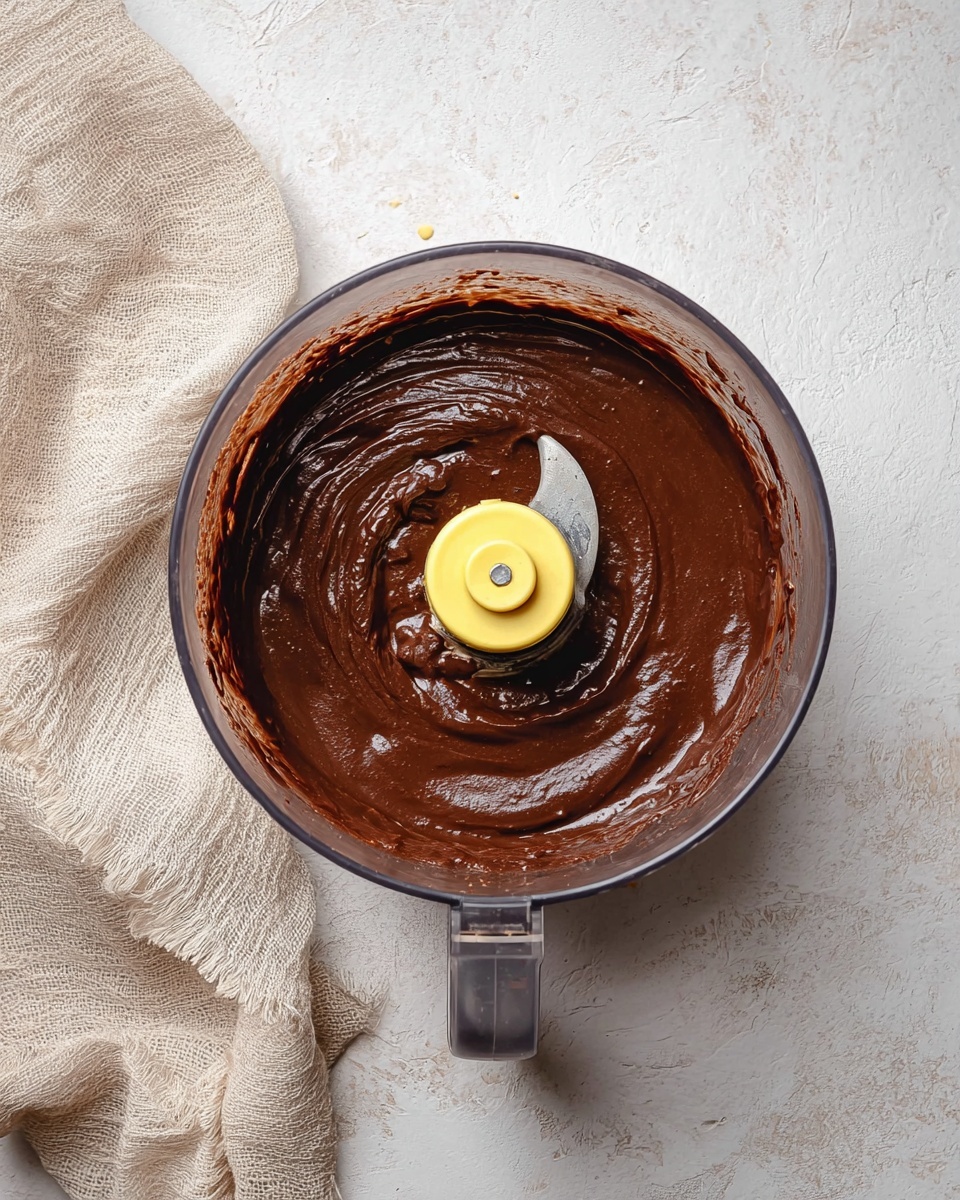 A clear food processor bowl filled with smooth, shiny dark brown chocolate batter that covers the bottom and sides evenly, with the yellow plastic blade attachment visible in the center. The bowl sits on a white marbled textured surface with a light beige linen cloth draped nearby. The image is captured from above, showing the thick, creamy texture of the chocolate mixture inside the bowl. photo taken with an iphone --ar 4:5 --v 7