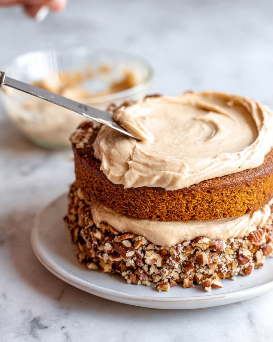 The image shows a two-layer cake on a white plate on a white marbled surface. Each layer is a brown cake sprinkled with chopped nuts on top, giving a rough texture. Between the layers and on top, there is a smooth light brown cream being spread with a silver knife. The cream looks thick and creamy, covering the cake partly. In the background, there is a glass bowl with more cream inside. A woman's hand is holding the knife spreading the cream. Photo taken with an iphone --ar 4:5 --v 7