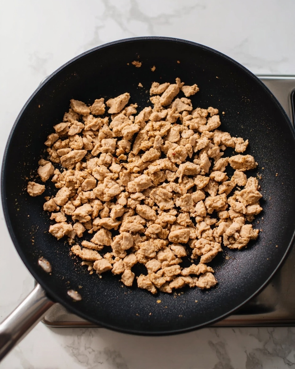 In the image, a black frying pan is shown from above, filled with small, light brown cooked meat pieces scattered evenly inside. The pan's matte black surface contrasts with the small chunks of meat that look soft and slightly crumbly. The pan rests on a stove with white elements partially visible around it. The white marbled surface below the stove adds a clean, bright feel to the scene. Photo taken with an iphone --ar 4:5 --v 7