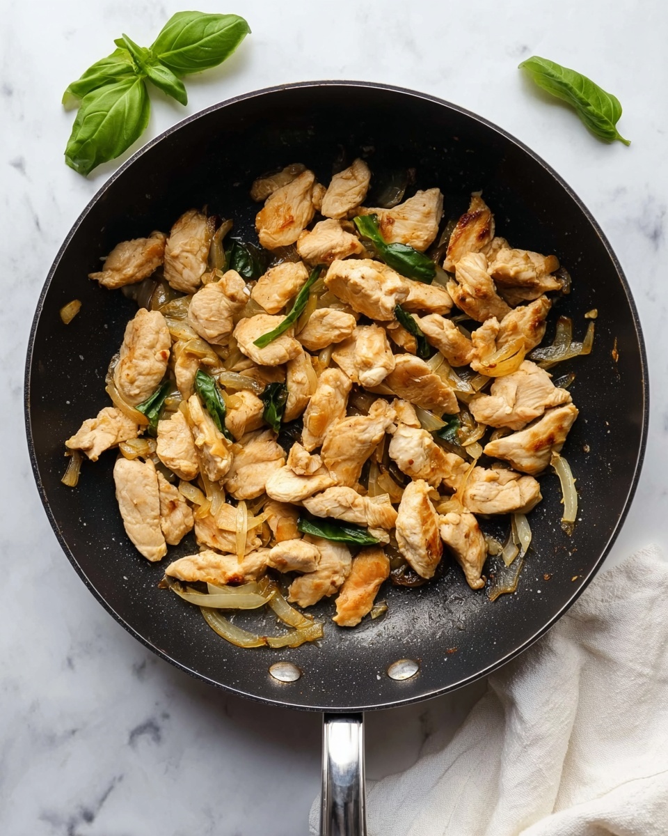 The image shows a black pan filled with cooked chicken pieces that are light golden brown and slightly crispy on the edges, scattered unevenly across the pan. Mixed in with the chicken are thin slices of translucent cooked onions, some slightly caramelized. The pan is placed on a white marbled surface with a few fresh green basil leaves nearby and a white cloth partially under the pan handle. The lighting highlights the texture of the chicken and onions, making them look tender and juicy photo taken with an iphone --ar 4:5 --v 7