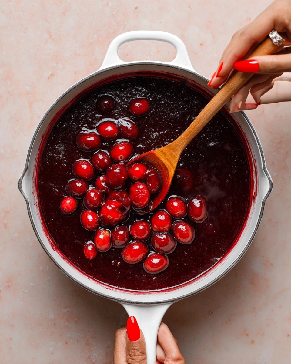 A white rectangular baking dish holds a stack of six dark brown glazed chicken wings with charred spots and a shiny texture, soaked in a deep red sauce mixed with whole red cranberries and sprigs of thyme. The sauce pools at the bottom, creating a rich, glossy layer under the wings. Around the dish, on a white marbled textured surface, are scattered fresh red cranberries, green herb sprouts, a split open pomegranate showing its bright red seeds, and a white plate brimmed with small red fruits with green leaves. Two glasses filled with ice and a dark drink are visible near the dish, along with golden forks placed on the sides. The photo taken with an iphone --ar 4:5 --v 7