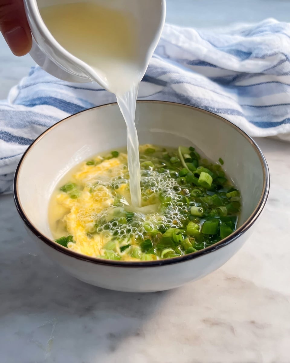 A white bowl with a thin dark rim sits on a white marbled surface, containing two layers: the bottom layer is a mixture of light yellow scrambled eggs and bright green chopped spring onions covering half the bowl. A clear liquid is being poured into the bowl from a small white cup held by a woman's hand, creating bubbles and a splash where it meets the eggs and onions. A soft, white cloth with blue stripes is in the background, slightly out of focus. photo taken with an iphone --ar 4:5 --v 7