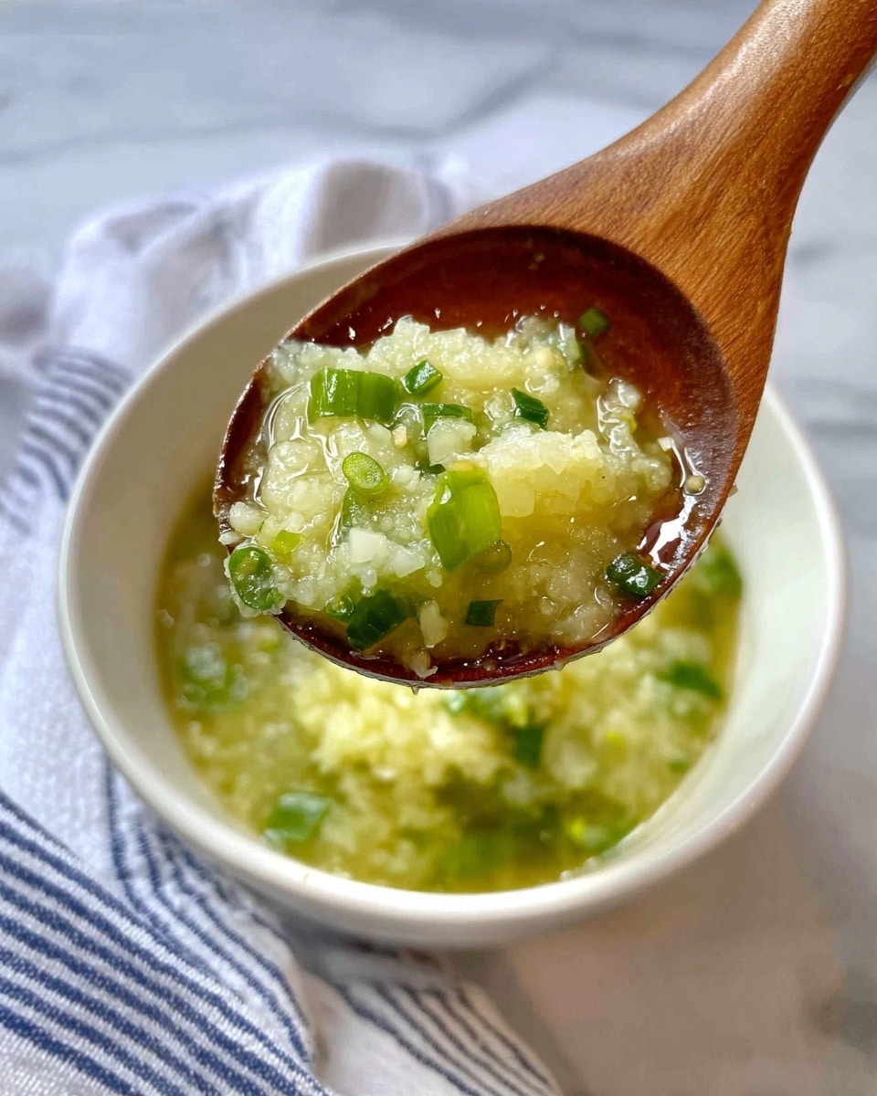 A close-up image shows a piece of light golden-brown cooked fish held by wooden chopsticks above a white bowl filled with green and yellow chunky dipping sauce mixed with herbs and oil. The fish has small green herb pieces on top, and the bowl sits on a white marbled surface with a few green herb pieces scattered around. In the background, a white cloth with dark stripes is slightly out of focus. photo taken with an iphone --ar 4:5 --v 7