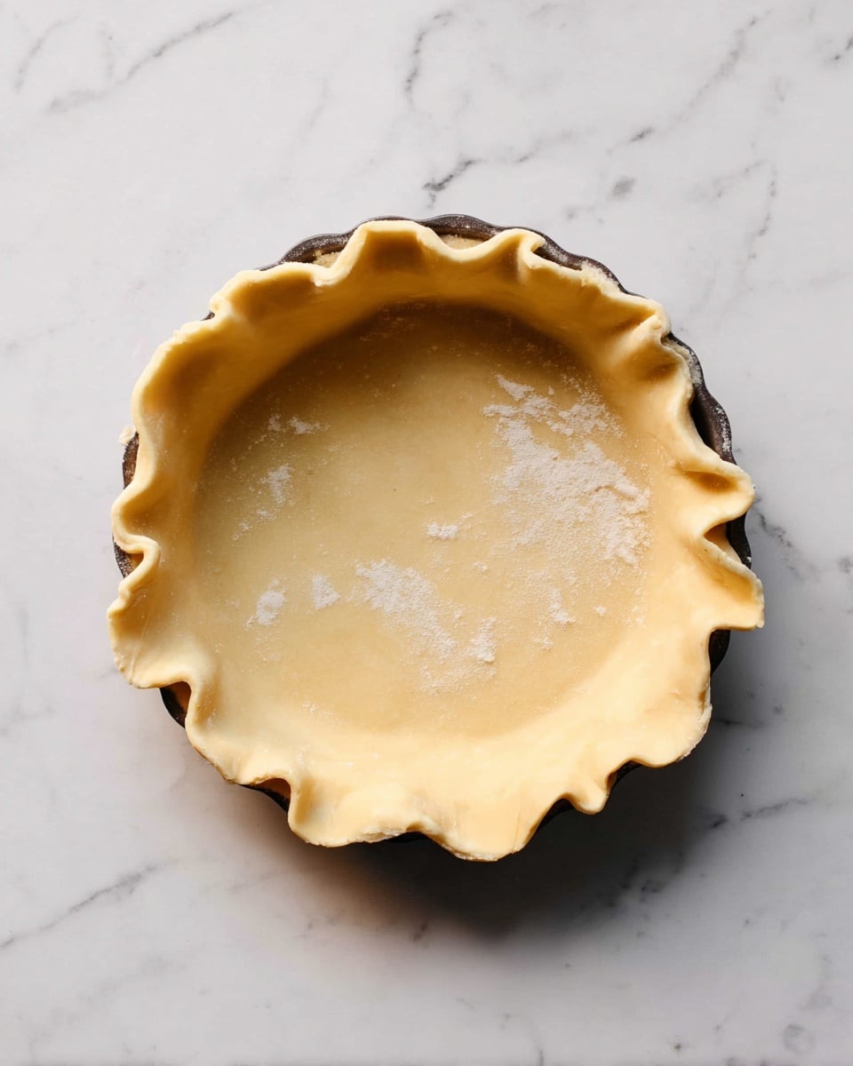 A round pie crust with a pale golden color is placed in a dark baking pan on a white marbled surface. The crust has an uneven edge that is folded and pinched in a wavy pattern around the rim, showing a soft, slightly flour-dusted texture. The inside of the crust is smooth and flat, ready for filling. Photo taken with an iphone --ar 4:5 --v 7