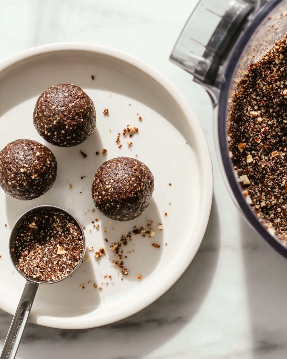 The image shows four round dark brown energy balls with a coarse texture, sprinkled with small light nut pieces on top, placed evenly on a white plate. To the left of the balls, there is a metal scoop with some energy mixture inside, resting on the plate. To the right side of the plate, part of a clear food processor bowl filled with a dark brown mixture with light nut bits is visible, set on a white marbled surface. The light is soft and natural, highlighting the texture of the balls and nuts. Photo taken with an iphone --ar 4:5 --v 7