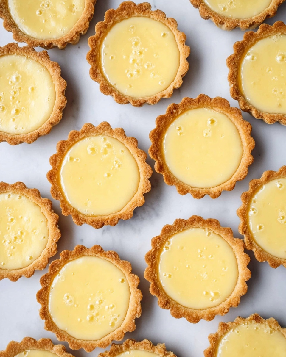 The image shows a small tart with two layers on a white plate with brown speckles, resting on a white marbled surface. The bottom layer is a smooth, yellow custard filling with a soft texture. The top layer is a fluffy white meringue with light brown toasty spots, shaped into a rounded peak with a swirl on top. One tart is cut in half, showing the cross-section clearly in the front, while more whole tarts are blurred in the background. A silver fork lies beside the cut tart. Photo taken with an iphone --ar 4:5 --v 7