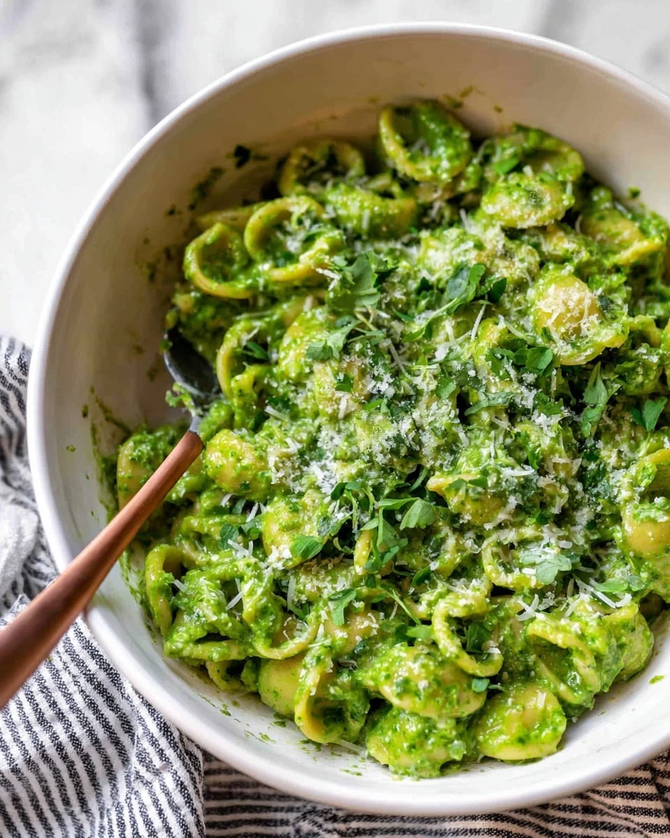 A white deep bowl is filled with short curly pasta covered in a thick, bright green sauce that looks creamy and smooth. On top of the pasta, there are small sprinkles of grated white cheese and some chopped fresh green herbs. A metal spoon with a long, thin, copper-colored handle is resting inside the bowl, partially covered by the pasta. The bowl is placed on a white marbled surface with a striped cloth nearby. photo taken with an iphone --ar 4:5 --v 7