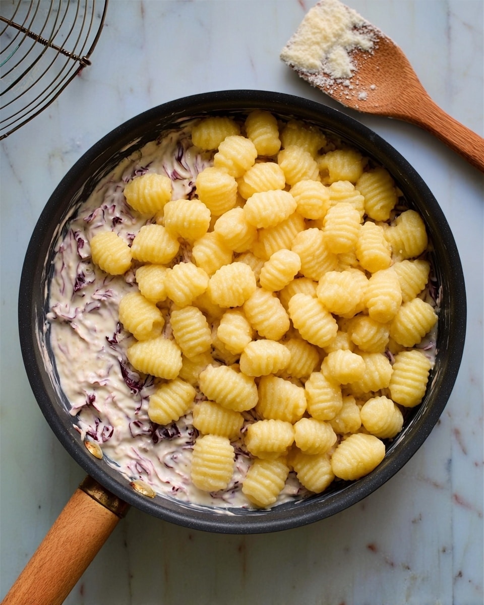 The dish is served in a white ceramic bowl filled with small, soft, round gnocchi pieces coated in a pale pink creamy sauce. Thin purple-red strips of radicchio are spread evenly throughout the sauce, adding a contrast of color and texture. The bowl sits on a dark brown cloth, which rests on a white marbled tabletop. Next to the bowl, on the right, are a fork and knife with silver handles. In the background, there is a stack of white plates with speckled edges, and a clear wine glass half-full of dark red wine on the right side. Photo taken with an iphone --ar 4:5 --v 7