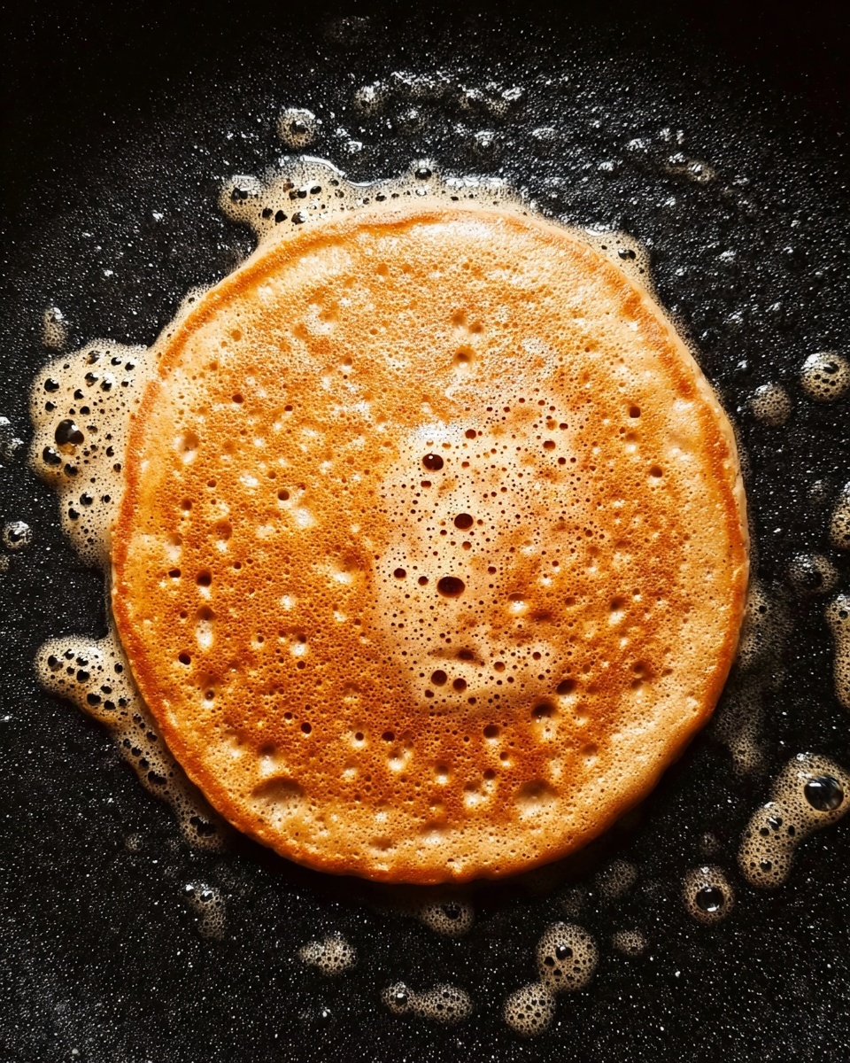 A single round pancake is shown cooking on a black non-stick pan with bubbles of melted butter around it. The pancake is golden brown with a slightly darker rim, and it has small bubbles and holes spread across its surface. The texture looks soft and slightly porous, with an even color showing light and darker brown spots. The background is the black pan's surface dotted with shiny, foamy butter spots. photo taken with an iphone --ar 4:5 --v 7