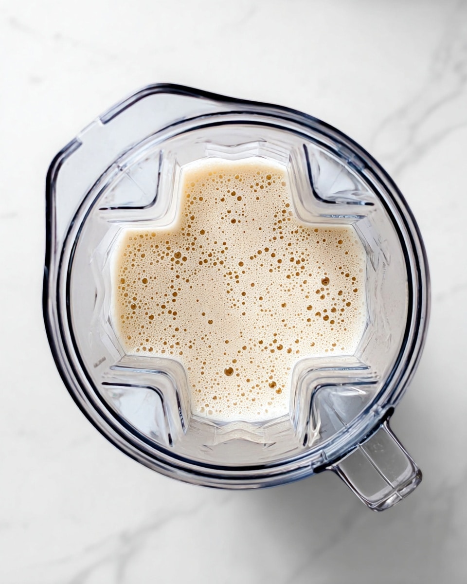 A clear blender jar is shown from above, filled about one-third with a light beige frothy liquid that has many small bubbles on its surface. The blender jar has a handle on the right side and a textured internal blade system visible inside. The jar sits on a white marbled surface that gives a clean and bright look. photo taken with an iphone --ar 4:5 --v 7