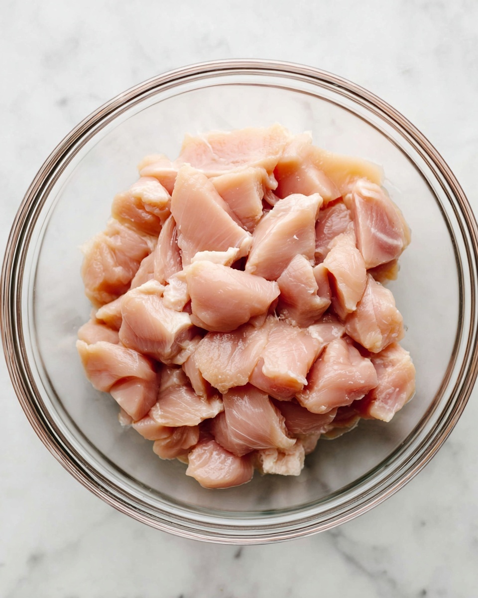 A clear glass bowl filled with raw chicken pieces, arranged in a loose pile. The chicken chunks are pale pink with slight variations in shade, showing smooth and slightly shiny textures. The bowl rests on a white marbled surface that adds a clean, fresh look to the scene. photo taken with an iphone --ar 4:5 --v 7
