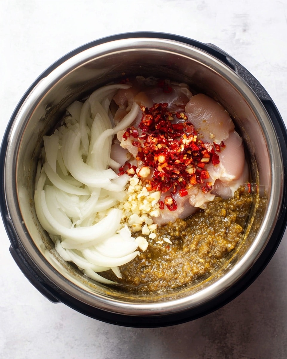 Inside a shiny silver pot, there is a base layer of thick green sauce with a chunky texture on the right side. On top of the sauce, there is a pink raw chicken piece slightly covered with red chopped peppers scattered around it. On the left side, there is a pile of white sliced onions with smooth, curved layers. Small bits of chopped garlic are sprinkled in the middle of the pot, resting partly on the onions and the chicken. The pot sits on a white marbled surface. photo taken with an iphone --ar 4:5 --v 7