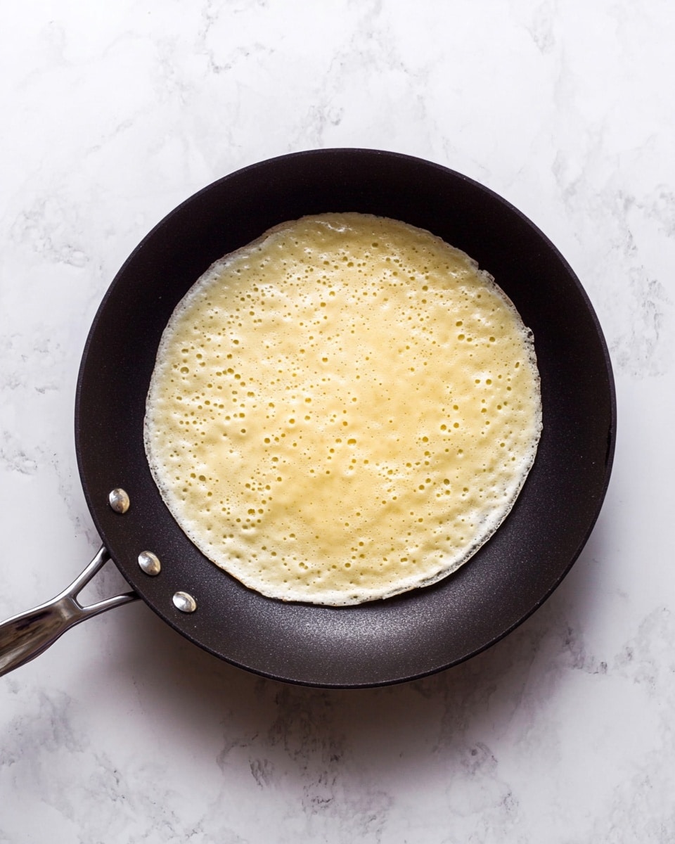 A single pancake is cooking in a black pan, positioned centrally. The pancake is light yellow with small bubbles and slightly darker edges, showing it is partially cooked. The pan has a simple, smooth texture with a dark matte finish and silver rivets attaching the handle. The background is a white marbled surface. photo taken with an iphone --ar 4:5 --v 7