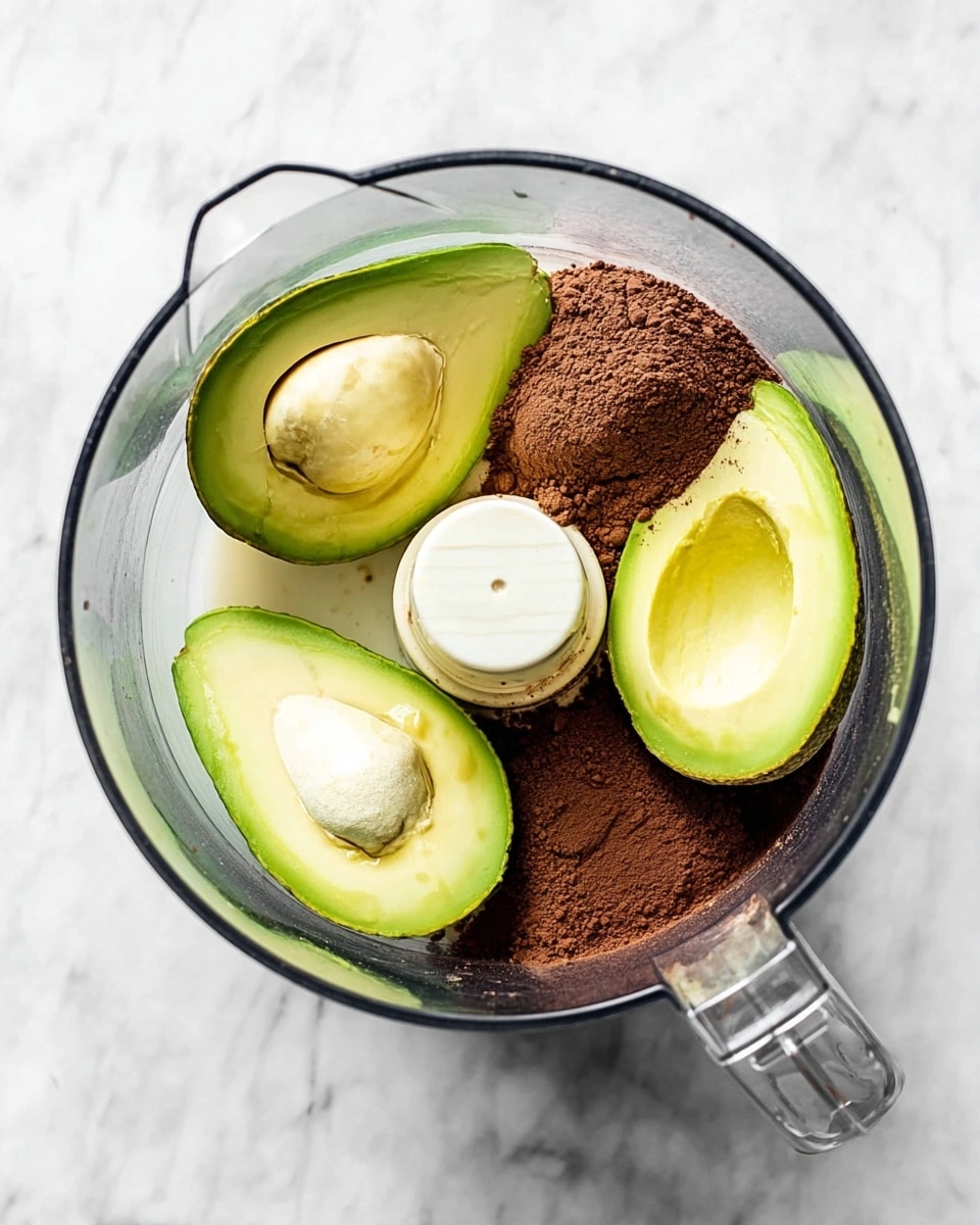 A clear glass food processor bowl sits on a white marbled surface, containing three avocado halves with smooth green skin and pale yellow-green flesh, and a pile of dark brown cocoa powder in one area. There is a small amount of light beige liquid pouring into the bowl from a glass container in the lower right corner. The food processor blade in the center is white, and the bowl edges are rounded and clear. The overall look is fresh with natural, creamy green avocados and rich cocoa powder. photo taken with an iphone --ar 4:5 --v 7