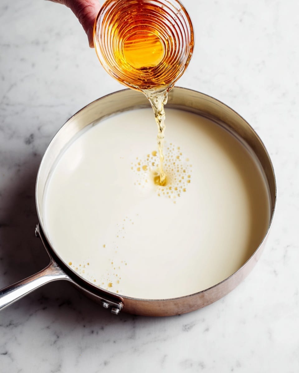 A silver pan filled with a smooth, creamy white liquid, with small bubbles around the edges. Above the pan, a woman's hand is pouring a golden amber liquid from a small ribbed glass cup into the white liquid. The scene is set on a white marbled surface, giving a clean and bright background. The light catches the glass and the liquid, showing a warm contrast between the two colors. photo taken with an iphone --ar 4:5 --v 7