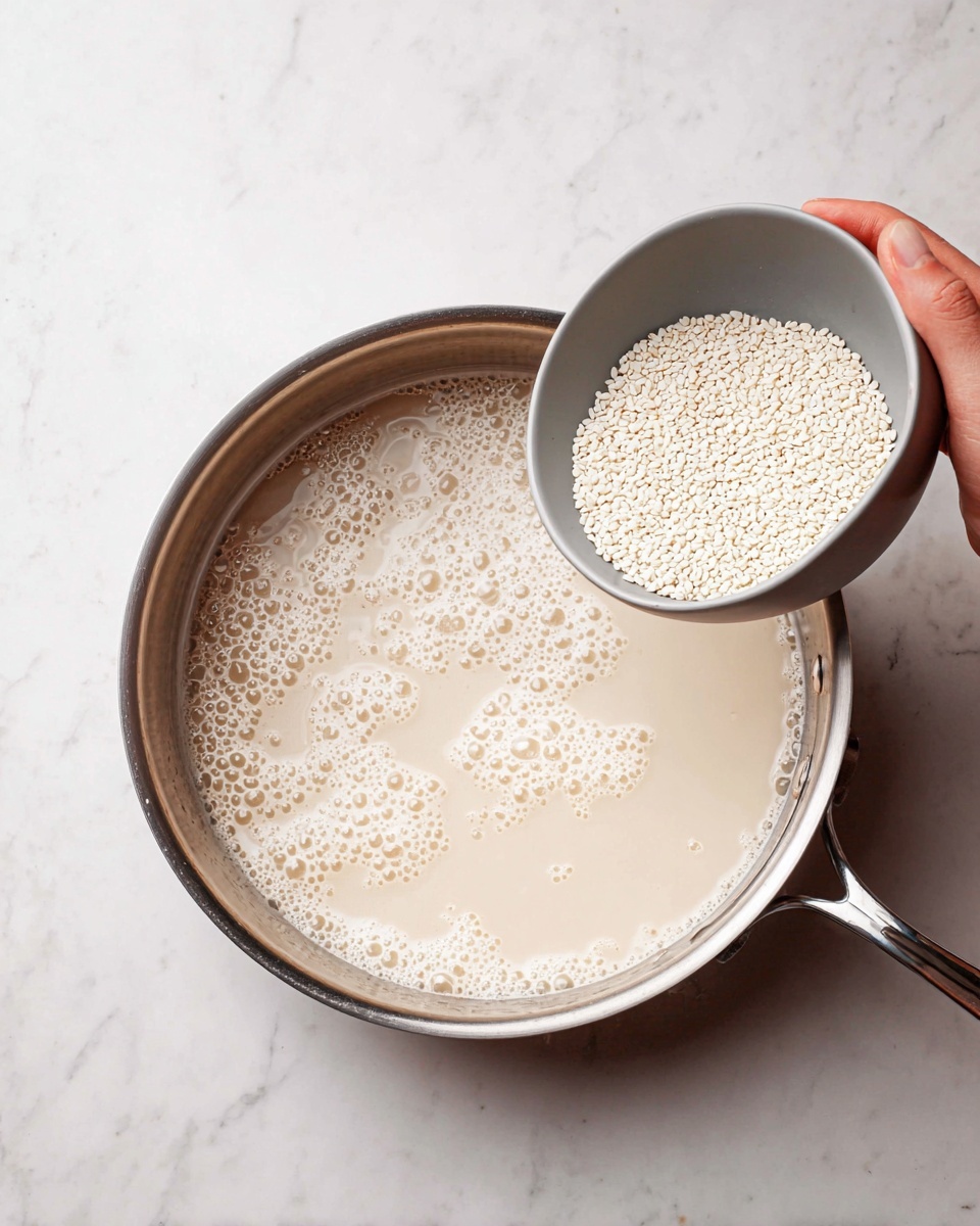 A close-up image shows a shiny metal pan filled with a light beige liquid with many small bubbles on its surface. A woman's hand is holding a small light gray bowl tilted above the pan, pouring small white round grains into the liquid. The pan is placed on a white marbled surface. photo taken with an iphone --ar 4:5 --v 7