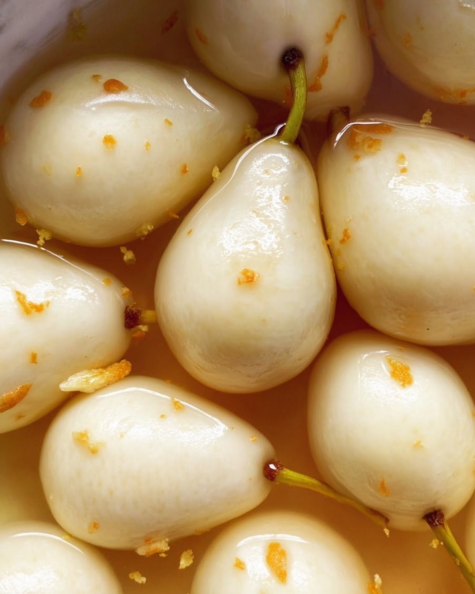 The image shows a close-up view of glossy, peeled white pears simmering in a light amber liquid with small bits of orange zest or spices scattered on the surface. The pears have a smooth texture with stems still attached on some. They are partially submerged and tightly packed, filling the frame from edge to edge, with the liquid reflecting soft light. The background has a white marbled texture. photo taken with an iphone --ar 4:5 --v 7