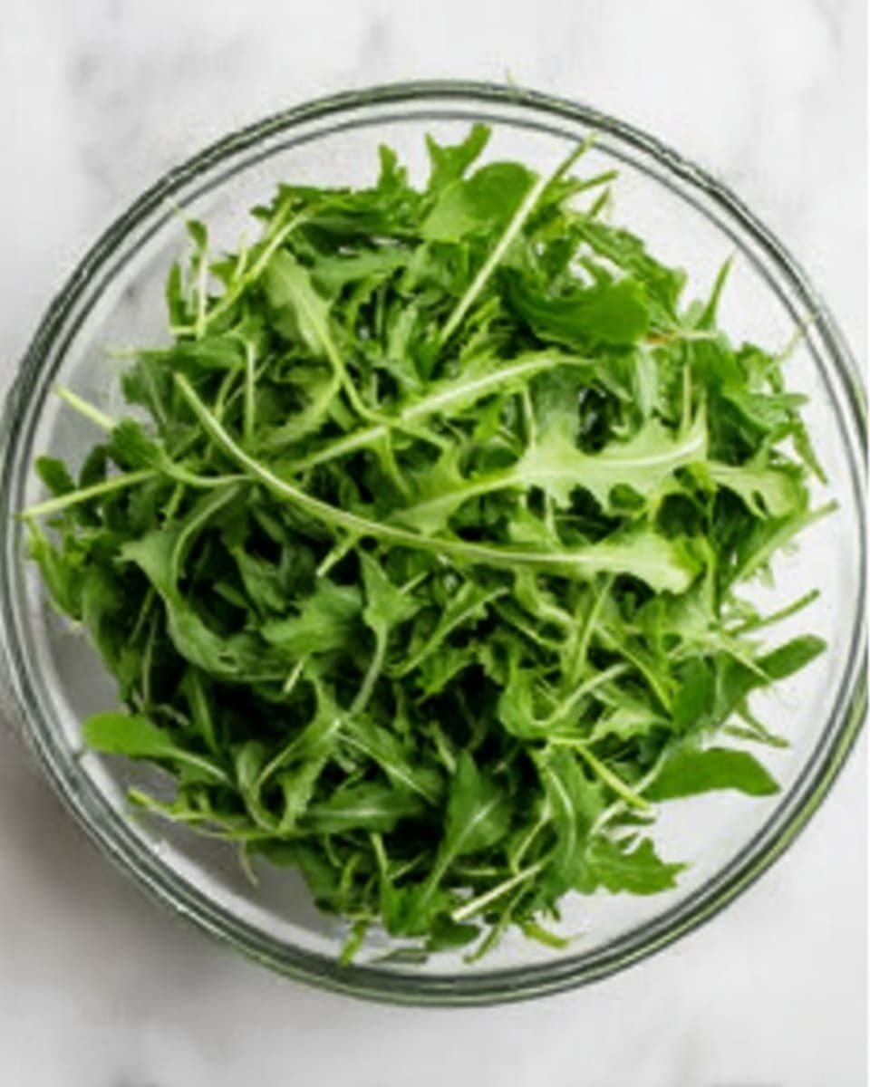 A clear glass bowl filled with bright green arugula leaves, piled high with a fresh, leafy texture. The arugula has small stems and slightly jagged edges, filling every corner of the bowl. The bowl is placed on a white marbled surface, giving a clean and fresh look. Photo taken with an iphone --ar 4:5 --v 7