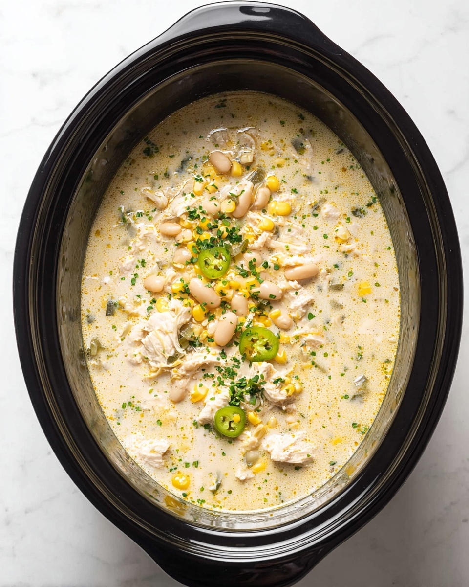 This image shows a white bowl filled with creamy chicken soup. The soup has a light greenish color and a smooth texture with small bubbles on the surface. Inside the soup, there are pieces of cooked chicken, white beans, and bits of yellow corn. On top, there are three golden brown, crispy tortilla strips arranged on one side. Two green chili slices and a sprig of fresh cilantro sit near the tortilla strips. A white dollop of sour cream or cream is also visible beside the tortilla strips. There are small green herbs sprinkled around the soup. The bowl sits on a white marbled surface. Photo taken with an iphone --ar 4:5 --v 7
