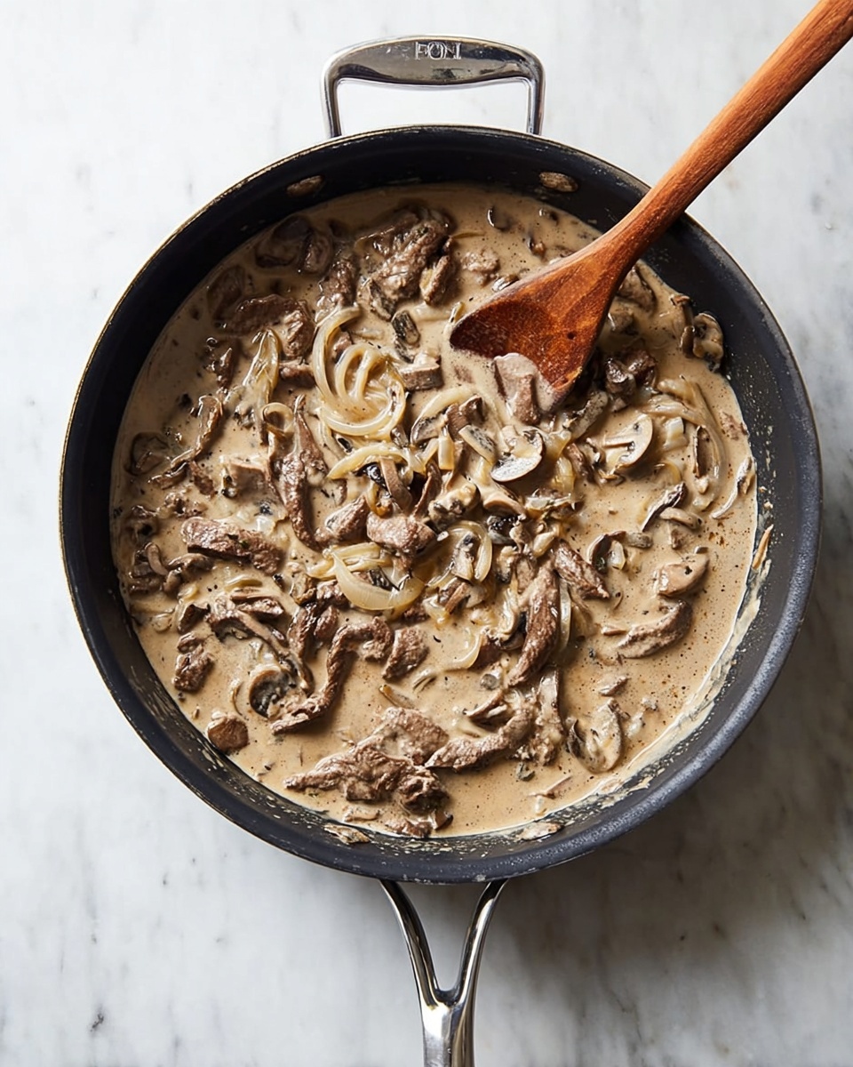 A black pan sits on a white marbled surface filled with a creamy sauce containing sliced mushrooms, thin strips of beef, and light-colored onion slices. A wooden spoon is dipped into the sauce on the right side of the pan. The creamy sauce has a light brown color, with visible textures of mushrooms and meat distributed evenly throughout. The pan has a shiny silver handle that extends downwards. photo taken with an iphone --ar 4:5 --v 7