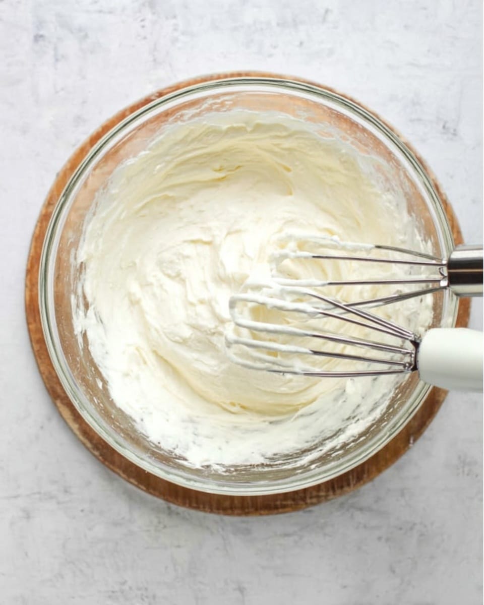 A clear glass bowl sits on a white marbled surface, filled with a thick white creamy mixture. Two metal beaters with white handles rest inside the bowl, covered lightly with the smooth, fluffy mixture. The texture looks soft and whipped, with small ridges showing the mixing motion. The overall scene is simple and clean, focused on the creamy content and the beaters. Photo taken with an iphone --ar 4:5 --v 7