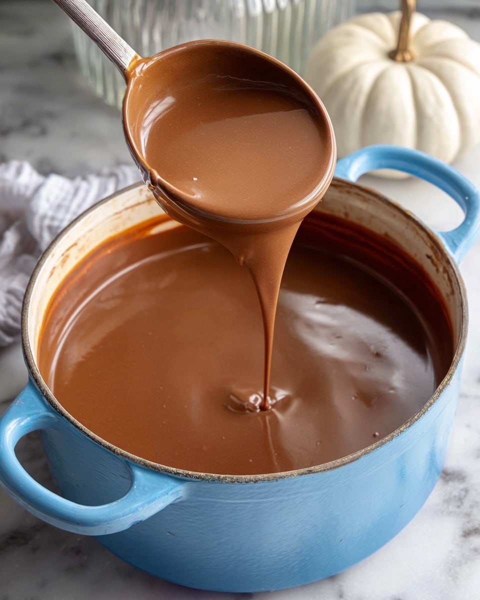 A close-up of a blue pot filled with smooth, shiny light brown chocolate sauce being stirred by a metal ladle coated in the same sauce, with the chocolate dripping back into the pot. The pot sits on a white marbled surface, and in the background, there is a white pumpkin and a ribbed glass jar adding subtle details to the scene. The sauce has a creamy texture with reflections showing a soft light source. Photo taken with an iphone --ar 4:5 --v 7