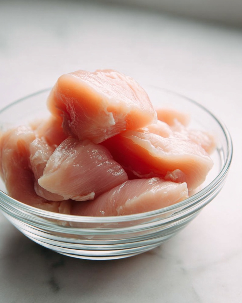 A clear glass bowl filled with several pieces of raw chicken, each piece pale pink and smooth with a slightly shiny texture, stacked loosely inside the bowl. The bowl sits on a white marbled surface with soft natural light highlighting the fresh and moist appearance of the chicken pieces. photo taken with an iphone --ar 4:5 --v 7