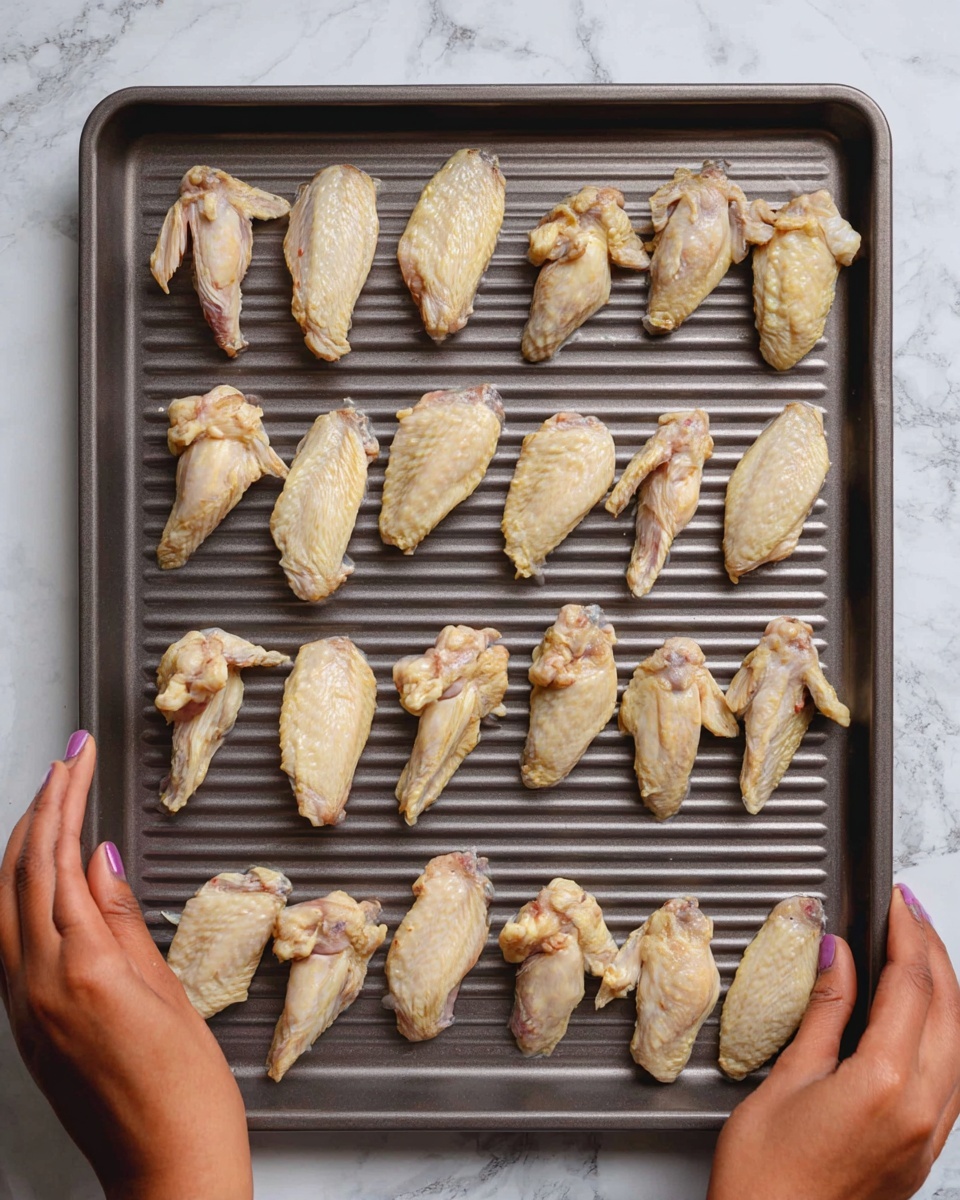 Sticky BlackBerry BBQ Wings Recipe 5 A dark gray metal baking tray with ridges holds eighteen pieces of raw chicken wings arranged in a grid, each wing showing pale yellow and light brown skin with visible bones, spaced evenly with tips pointing in different directions. Two woman's hands hold the tray on opposite sides, with visible darker skin tone and short nails. The background is a white marbled surface. photo taken with an iphone --ar 4:5 --v 7