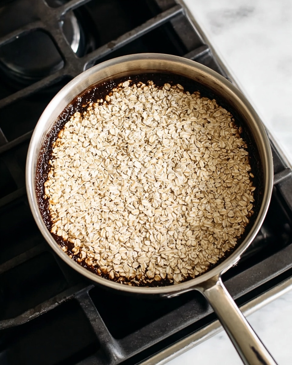 There are nine small, round dark brown cookie dough piles with visible oat pieces arranged on a baking tray lined with white parchment paper. To the left, a shiny silver pot holds more of the same cookie dough, thick and chunky with oats, with a silver spoon resting inside it. The tray and pot sit on a smooth white marbled surface. The scene is bright and clean with natural lighting. photo taken with an iphone --ar 4:5 --v 7