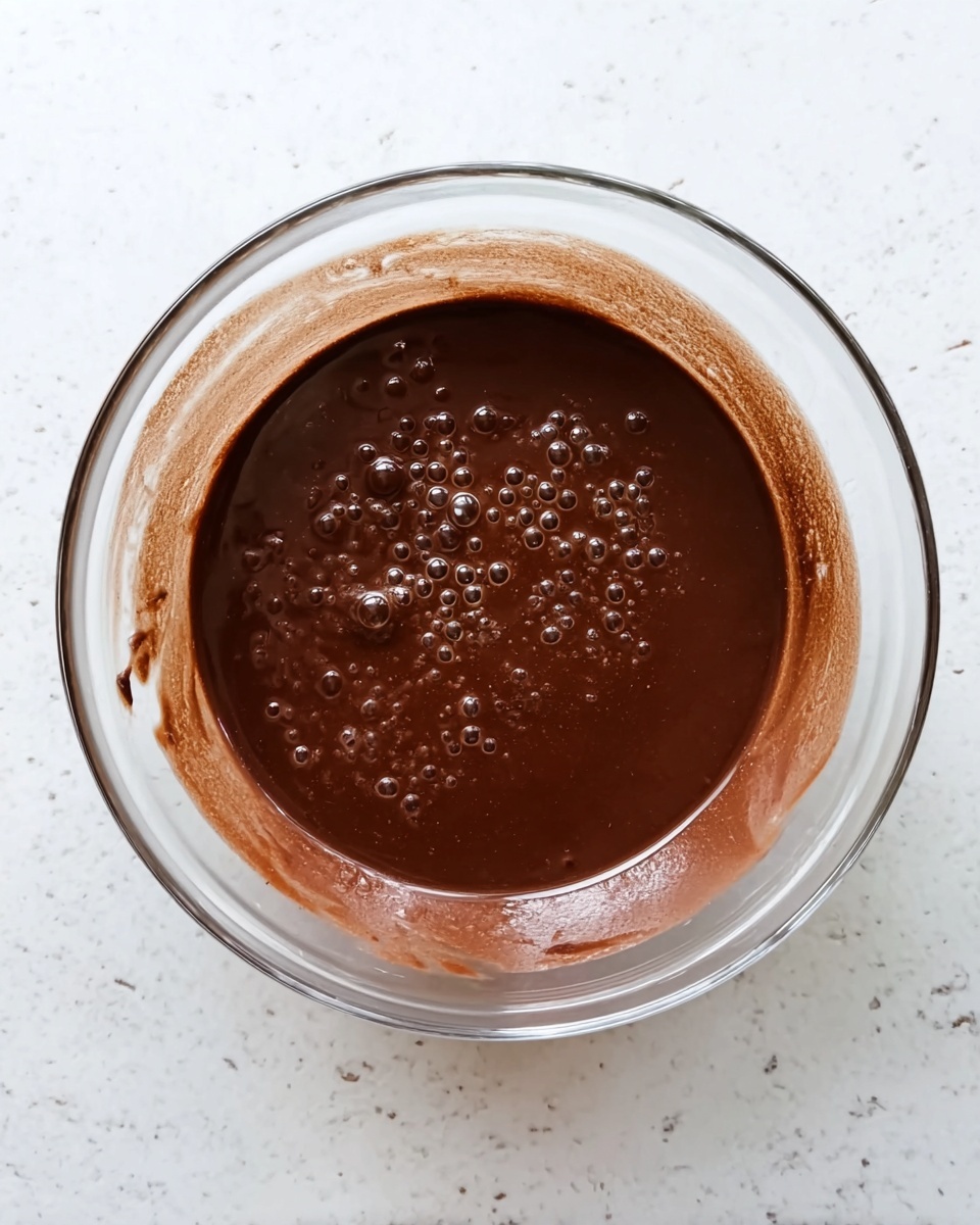 A clear glass bowl sits in the center filled with a smooth, dark brown chocolate mixture with small bubbles on the surface. The sides of the bowl have streaks of the chocolate mixture, showing the texture is thick but fluid. The bowl is placed on a white marbled surface, which gives a clean and bright background contrast to the rich, dark chocolate inside. photo taken with an iphone --ar 4:5 --v 7