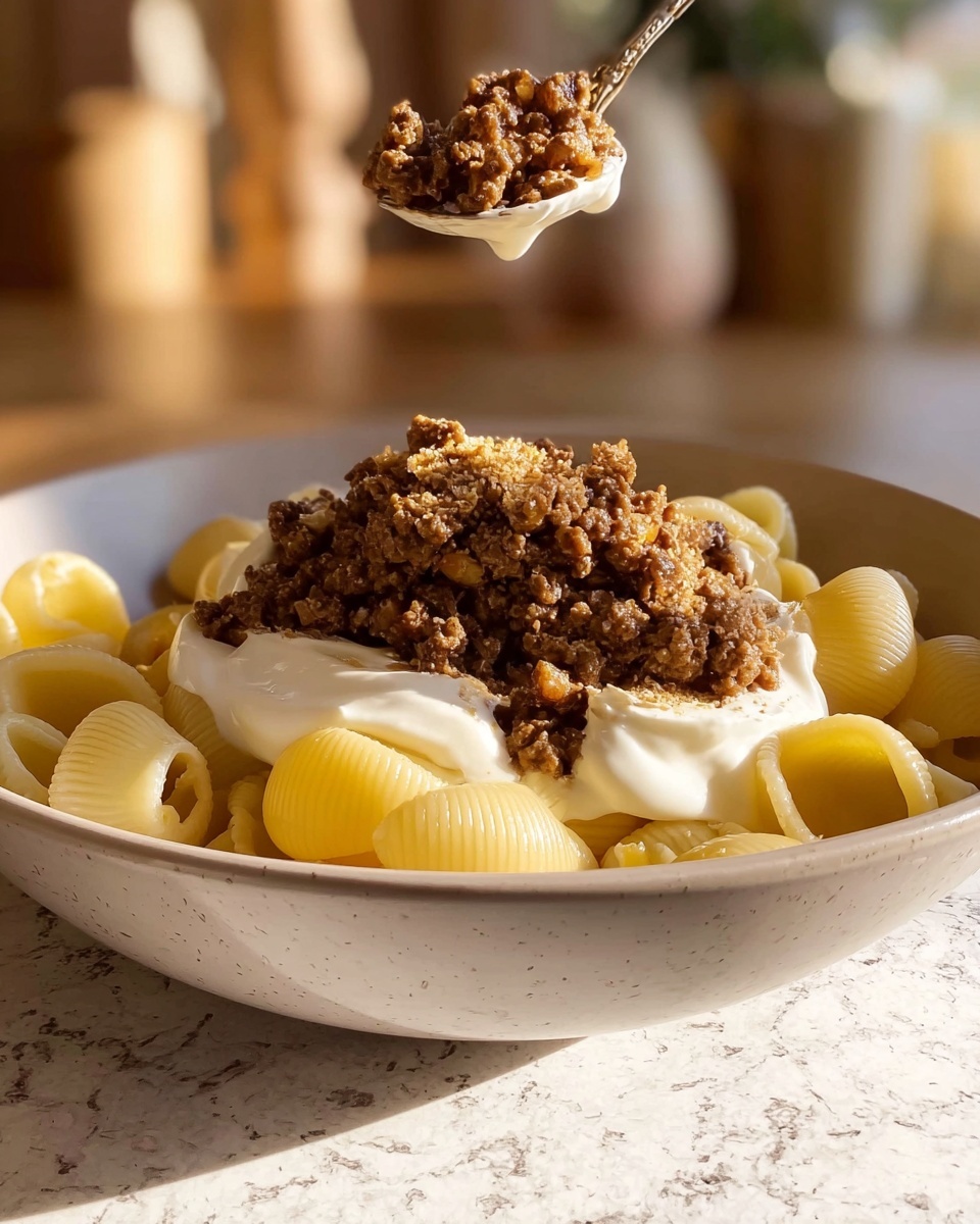 In a close-up shot, there is a white bowl filled with three main layers: the bottom layer consists of light yellow pasta shells with smooth, curved edges; on top of the pasta is a dollop of thick, creamy white sauce sitting in the middle; above the sauce, a spoon is placing a generous heap of finely crumbled cooked ground meat mixed with small pieces of cooked onions, creating a textured layer with brown and golden hues. The background features a warm, softly lit room with blurred details, and the whole scene is set on a surface with white marbled texture. Photo taken with an iphone --ar 4:5 --v 7