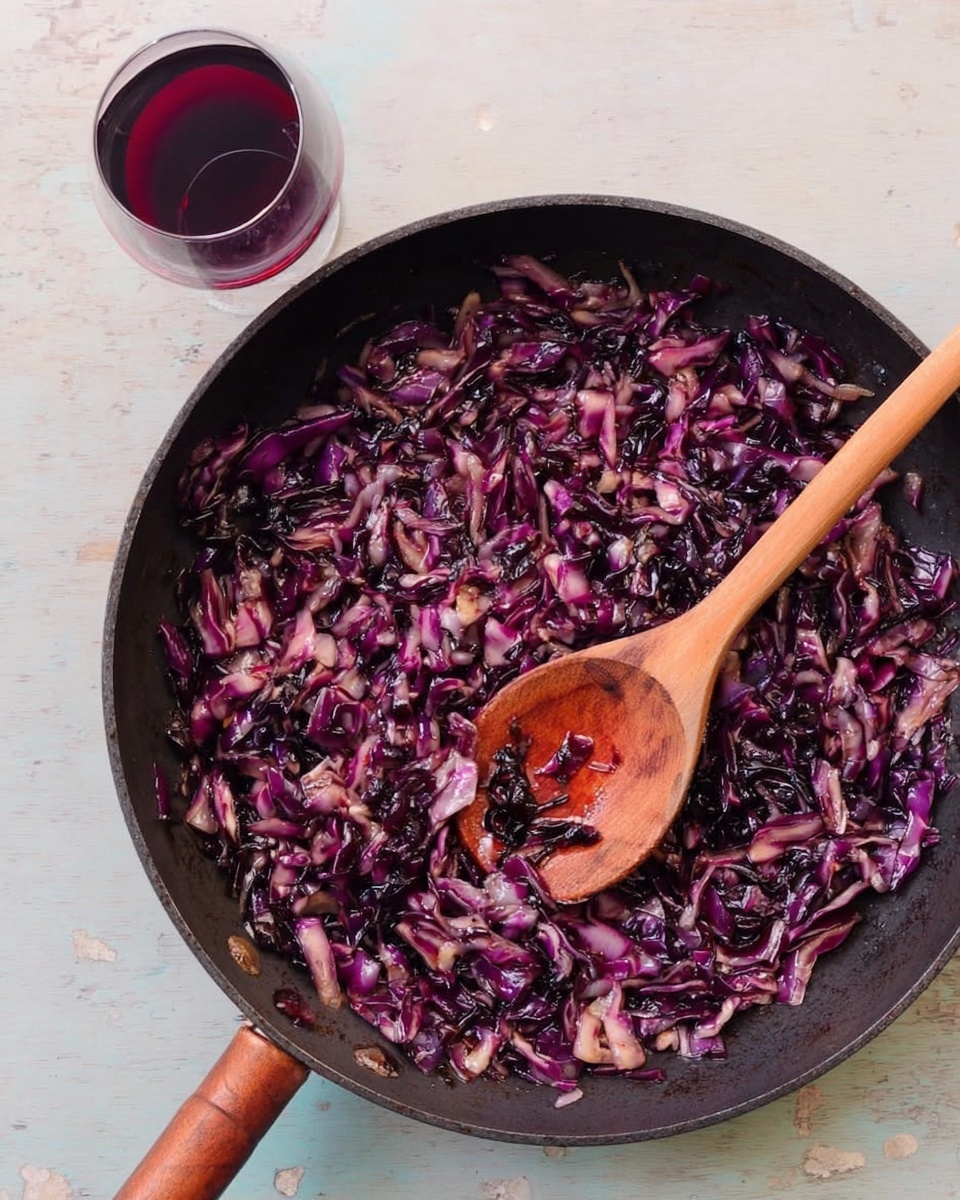 A black pan filled with finely chopped cooked red cabbage that looks soft and slightly shiny, with pieces varying in deep purple and light pinkish colors mixed evenly throughout. A wooden spoon with a light brown handle rests inside the pan, partly covered by the cabbage. Next to the pan is a clear wine glass holding a small amount of red wine. The pan is placed on a white marbled surface. photo taken with an iphone --ar 4:5 --v 7