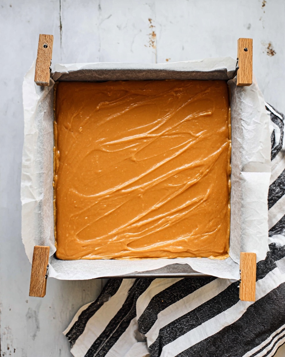 A stack of eight large, soft, square orange fudge pieces sits on a white plate. The fudge has a smooth, creamy texture with small air holes throughout and one piece on top has a small bite missing, showing its dense inside. The plate is placed on a white marbled surface, with a blurred background of a bottle and a dark cloth. A few small orange fudge bits are scattered near the plate. The overall colors include warm orange tones of the fudge contrasting with the clean white plate and surface. photo taken with an iphone --ar 4:5 --v 7