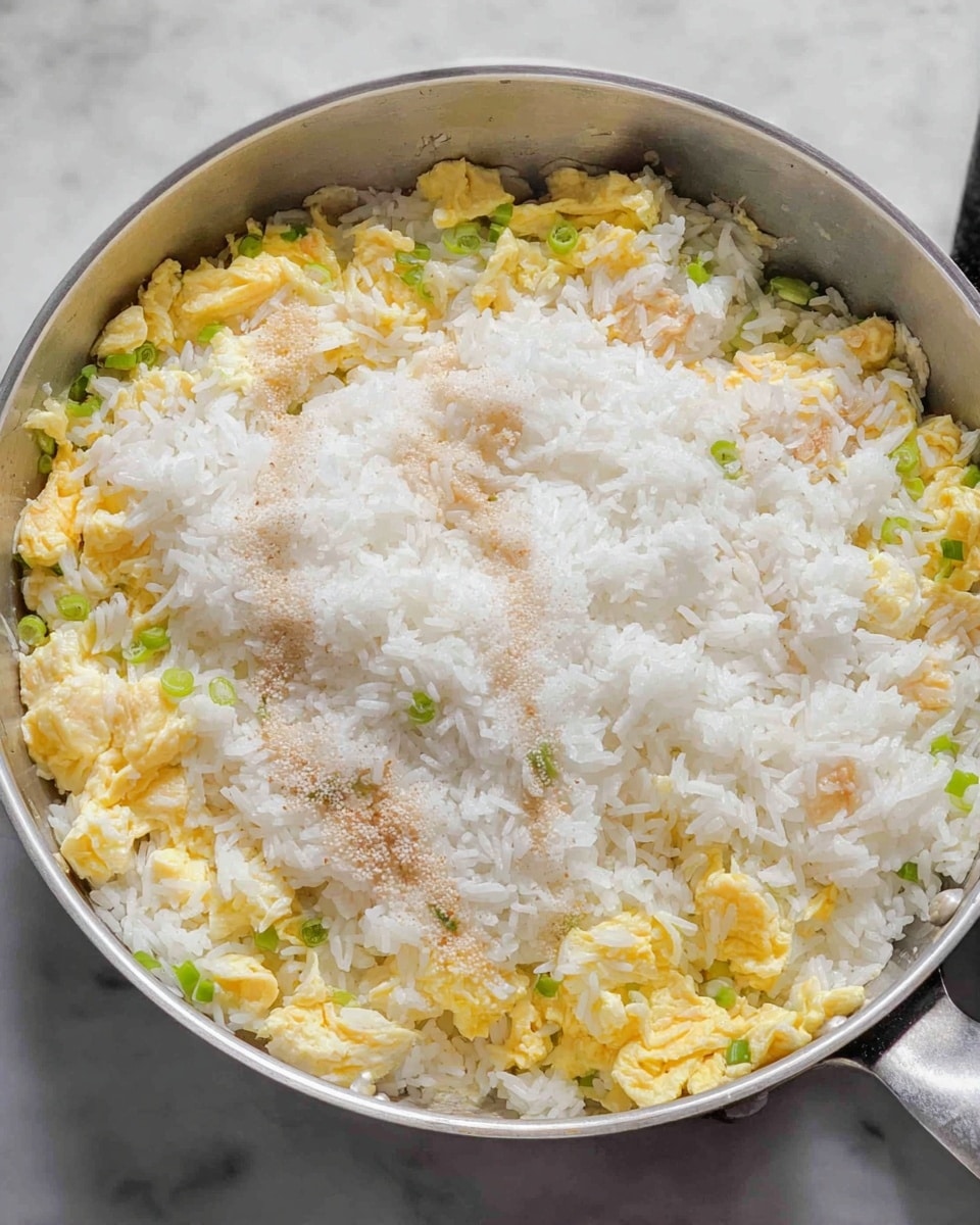 A close-up view of a frying pan filled with three visible layers: the bottom layer is scrambled eggs with light yellow color and small pieces of green onion, the middle layer is plain white rice covering the eggs, and the top layer shows sprinkled white granulated salt and a bit of light brown soy sauce spread unevenly over the rice. The frying pan is silver, and it sits on a white marbled surface. Photo taken with an iphone --ar 4:5 --v 7