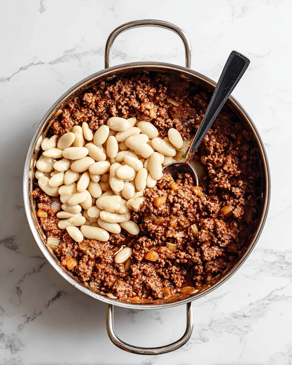 A metal pan with two handles sits on a white marbled surface, filled mostly with cooked ground meat mixed with small pieces of onion, showing a rich brown color and slightly oily texture. On top of the meat in the center is a pile of creamy white beans, smooth and slightly shiny. A black spoon with a metal handle is resting inside the pan, scooping some of the meat mixture near the edge of the beans. The pan and food are well-lit, highlighting the contrast between the dark meat and the pale beans. photo taken with an iphone --ar 4:5 --v 7