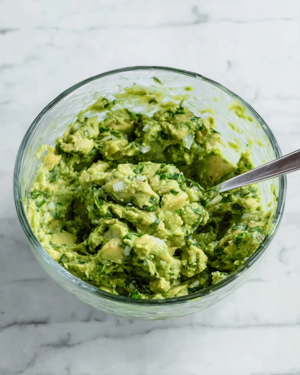 A clear glass bowl filled with a chunky green mix that looks like mashed avocado mixed with small pieces of herbs and onions, giving it a textured, fresh appearance. A silver spoon is resting inside the bowl on the right side, partially covered with the green mixture. The bowl sits on a white marbled surface that has subtle grey veins. The focus is on the thick, uneven texture of the green mix in the bowl. photo taken with an iphone --ar 4:5 --v 7