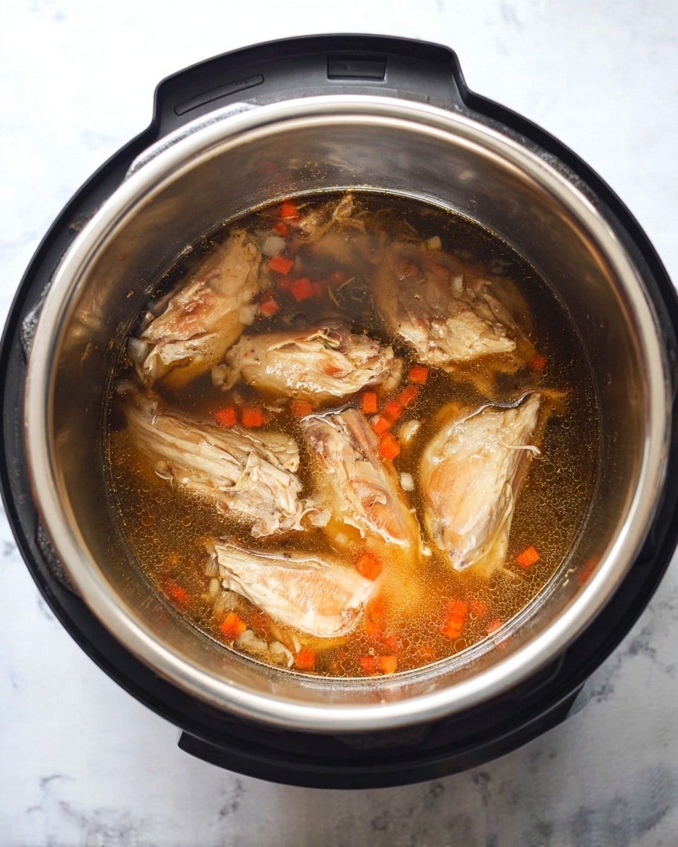 The image shows an Instant Pot with a shiny silver inner pot filled with a clear brown broth containing six large pieces of cooked chicken. The chicken pieces are light brown with visible strands and some slight red-brown char marks. Inside the broth, small diced orange carrot pieces and bits of onion float around. The Instant Pot sits on a white marbled surface, and the photo is taken from a bird’s-eye view, showing the contents clearly. photo taken with an iphone --ar 4:5 --v 7