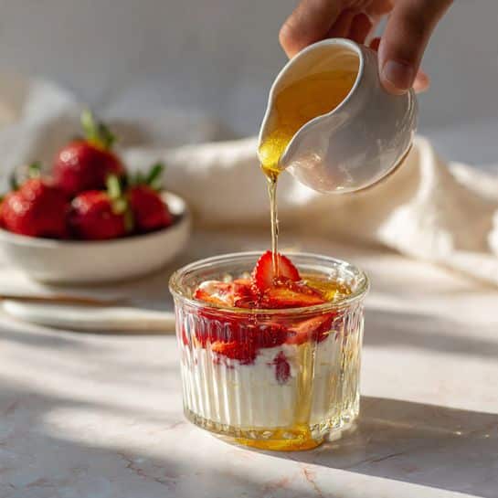 A clear glass with vertical ridges filled with white yogurt layered with small pieces of red strawberries inside. A woman's hand is pouring golden honey from a small white ceramic jug into the top of the yogurt, where two strawberry slices are visible. In the background, there is a white bowl filled with whole strawberries, sitting on a white marbled surface with a soft white cloth partly visible. The light is soft, highlighting the creamy texture of the yogurt and the shiny honey photo taken with an iphone --ar 4:5 --v 7